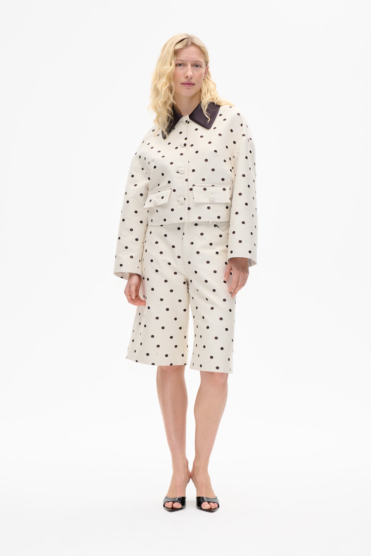 Model wearing a cream jacket with dark polka dots, oversized dark collar and flap pockets, paired with matching knee-length shorts.