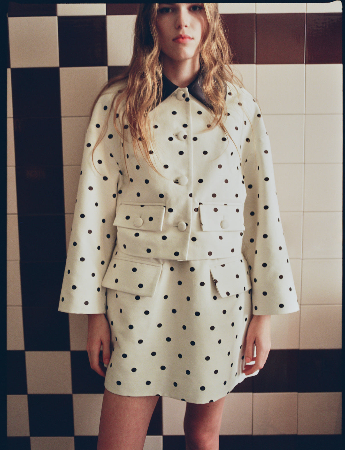 Model wearing a white jacket with black polka dots and large buttons, paired with a matching mini skirt against a checkerboard wall.