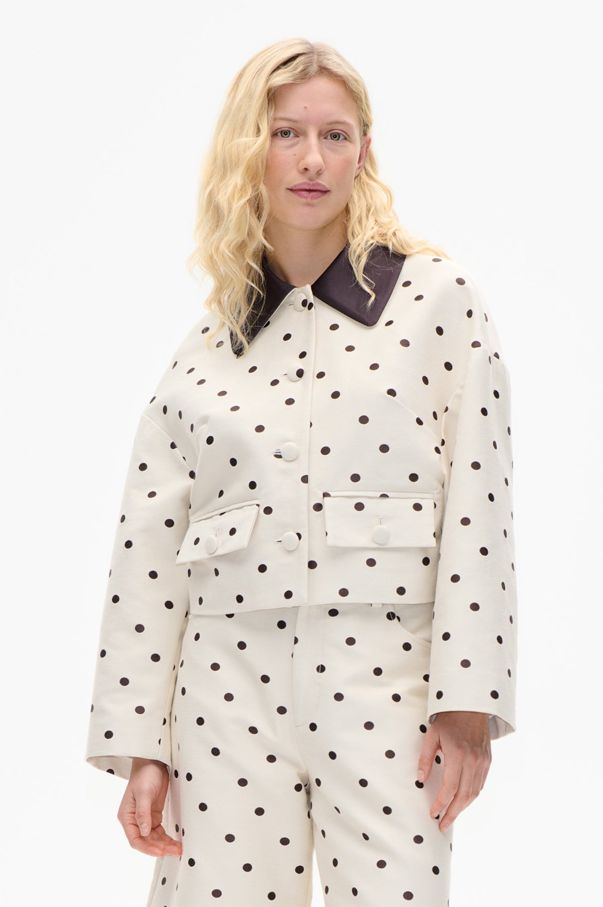 Model wearing a cream polka-dot coat with a dark contrasting collar, white buttons and flap pockets, paired with matching trousers.