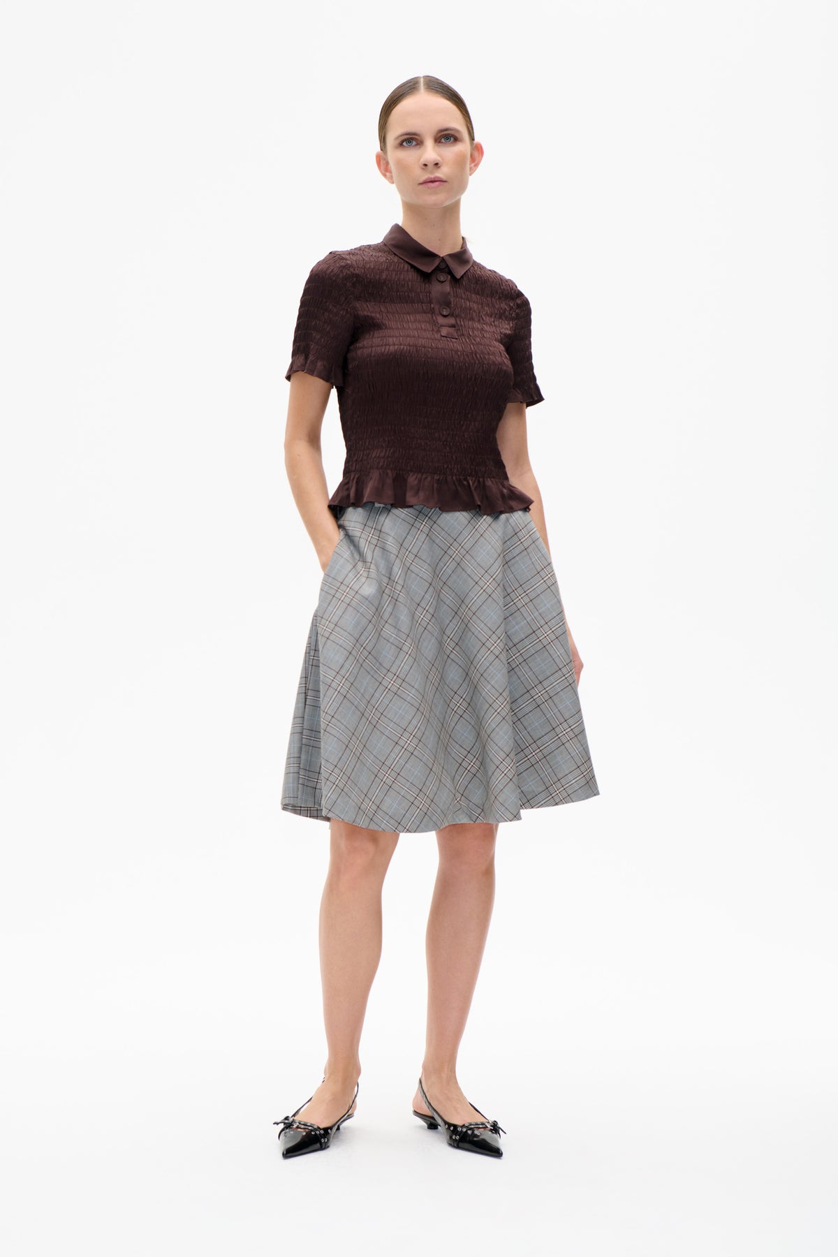 Model wearing a dark plum textured short-sleeve blouse with a rounded collar and peplum hem, paired with a grey plaid skirt.