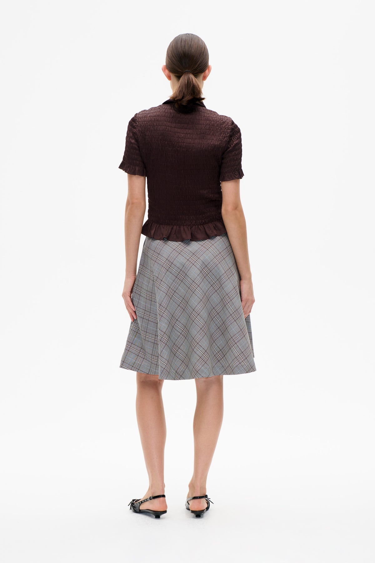 Back view of a model wearing a dark brown smocked blouse with short ruffled sleeves and a grey plaid skirt.