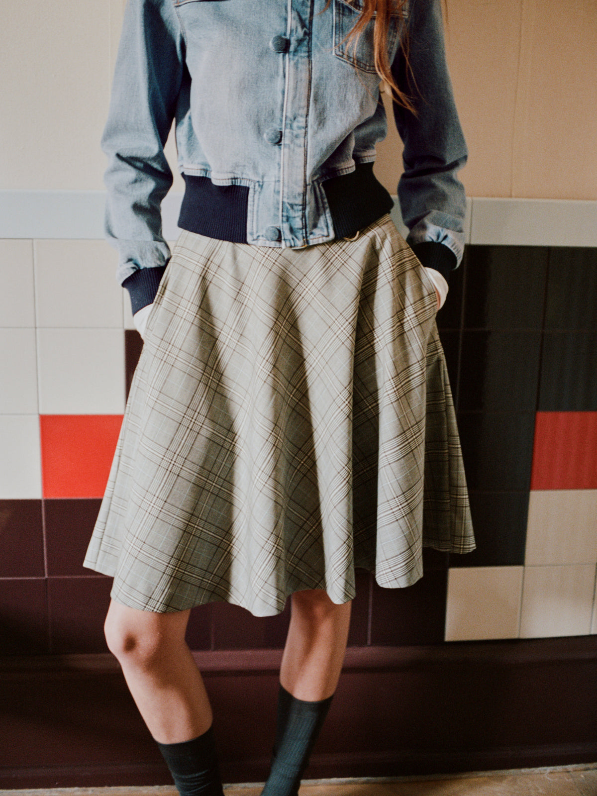 Gecropte campagnefoto van een persoon met een lichtblauwe denimjas en een beige geruite midirok tegen een betegelde muur.