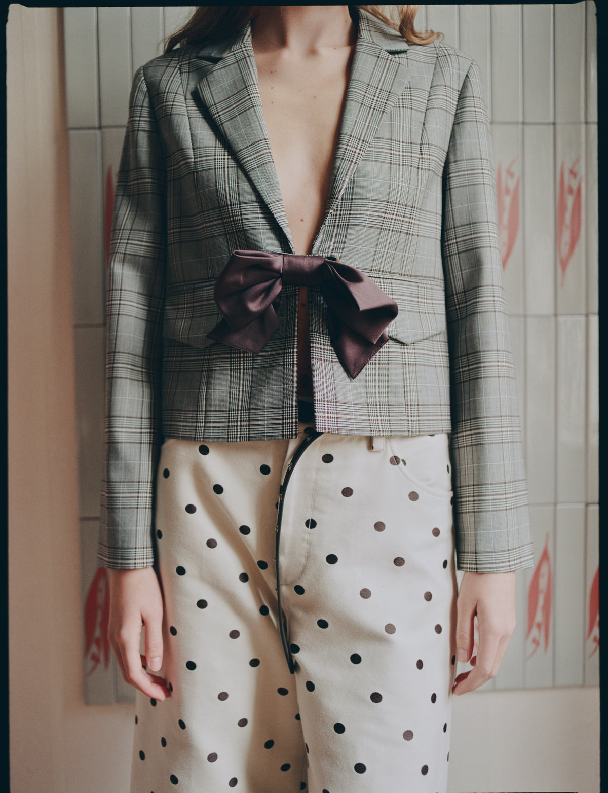 Close-up campaign view of a model wearing a grey plaid blazer with a burgundy bow and a cream skirt patterned with dark polka dots.