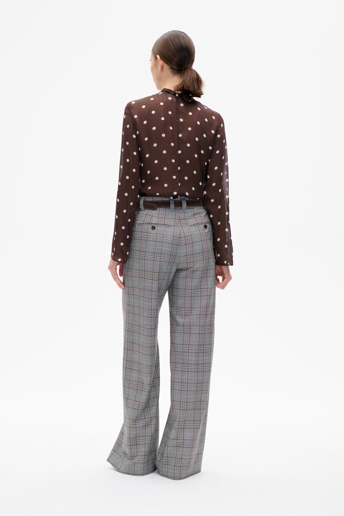 Back view of a model wearing a sheer brown blouse with white polka dots and grey plaid trousers with a belt.
