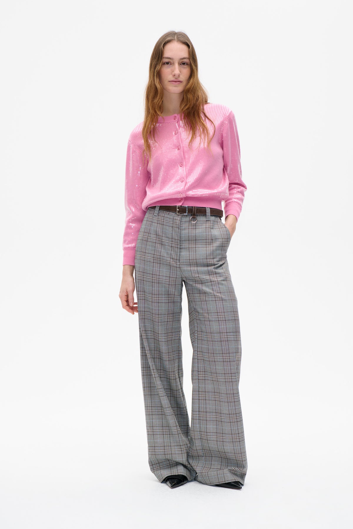 Model wears a pink button-front cardigan with white geometric sequin detailing and wide-leg grey plaid trousers, shown from the front.