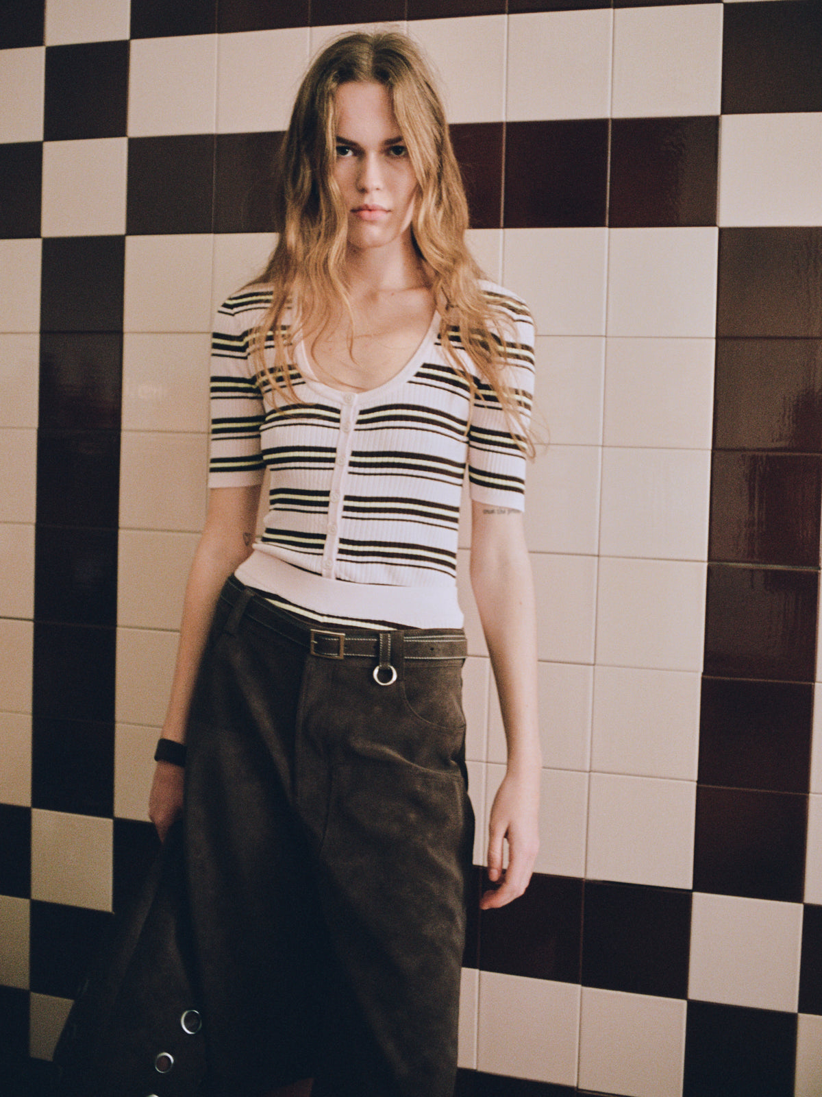 Model in een witte top met knoopsluiting en korte mouwen met zwarte strepen en een donkere broek, tegen een wand met zwart-witte dambordtegels.