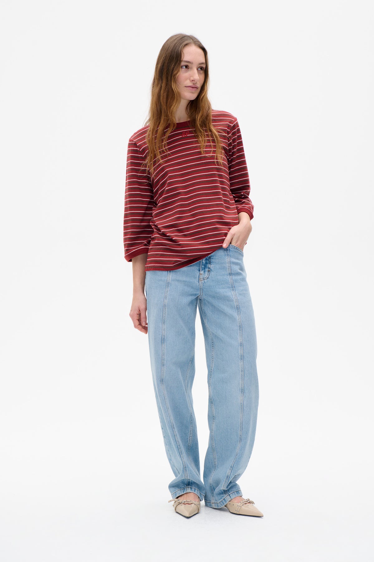 Model wearing a red long-sleeved top with thin horizontal stripes and a small chest logo, paired with light-wash jeans.