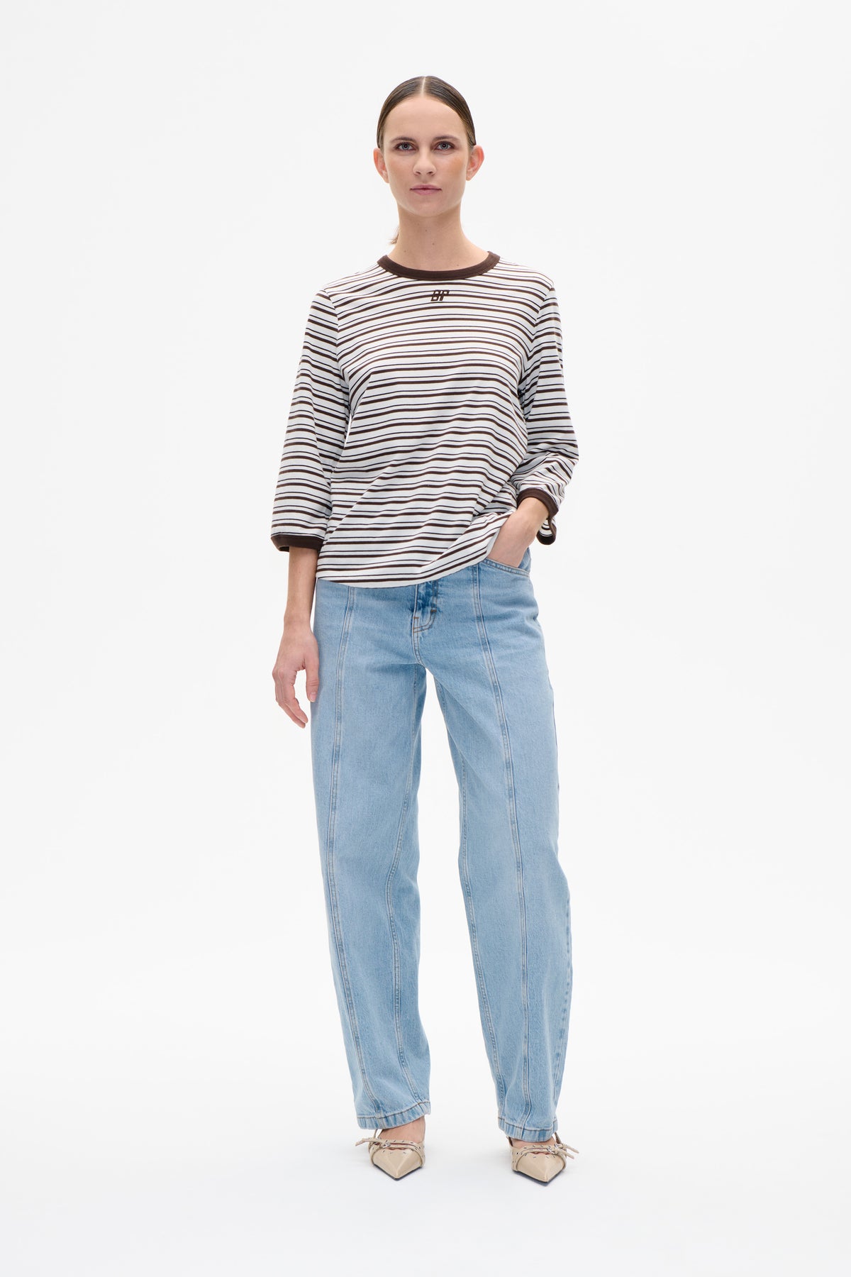 Front view of a model wearing a cream long-sleeved T-shirt with dark brown stripes, contrast trim and a small chest logo, paired with light-wash jeans.