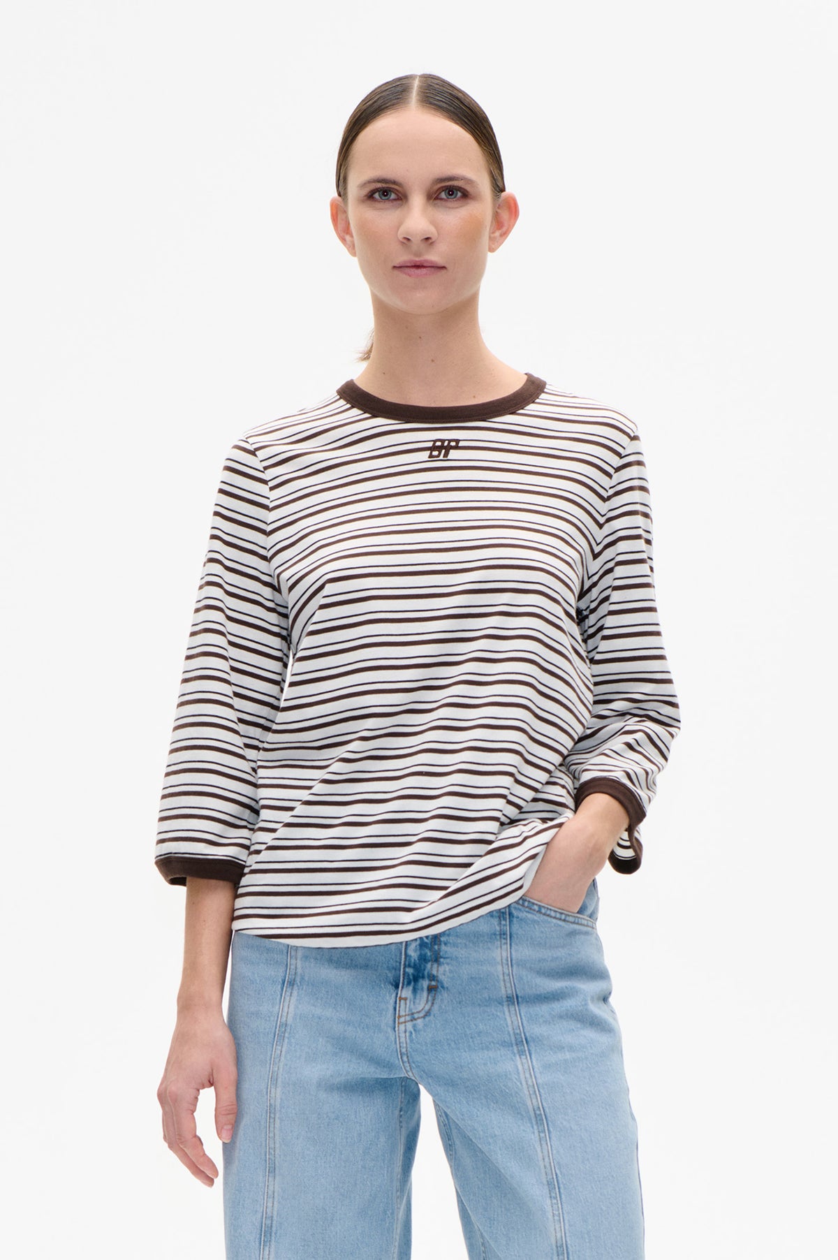Model wearing a white long-sleeved top with dark brown horizontal stripes, contrast trim and a small chest logo, paired with light blue jeans.