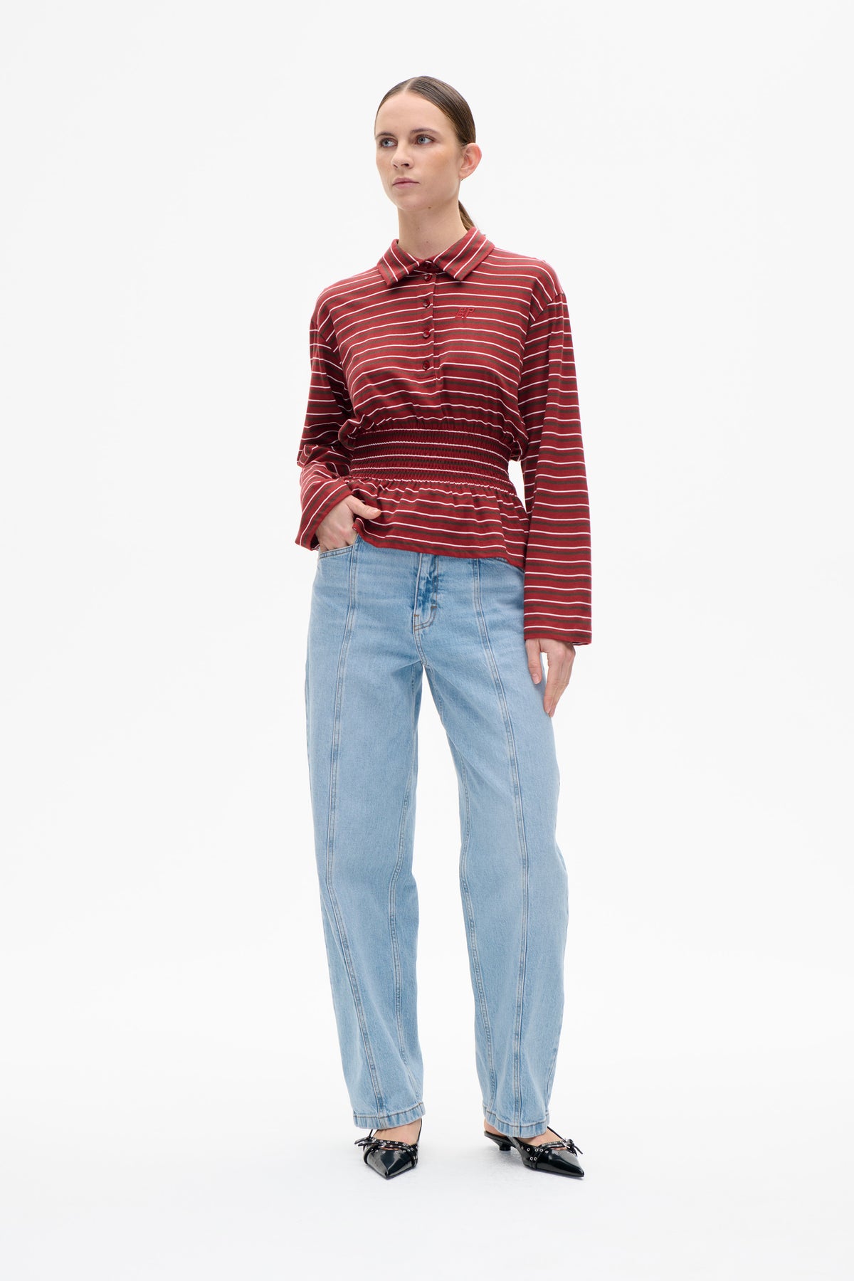 Model wearing a red and brown striped long-sleeve button-front top with a pointed collar and gathered waist, paired with light blue jeans.