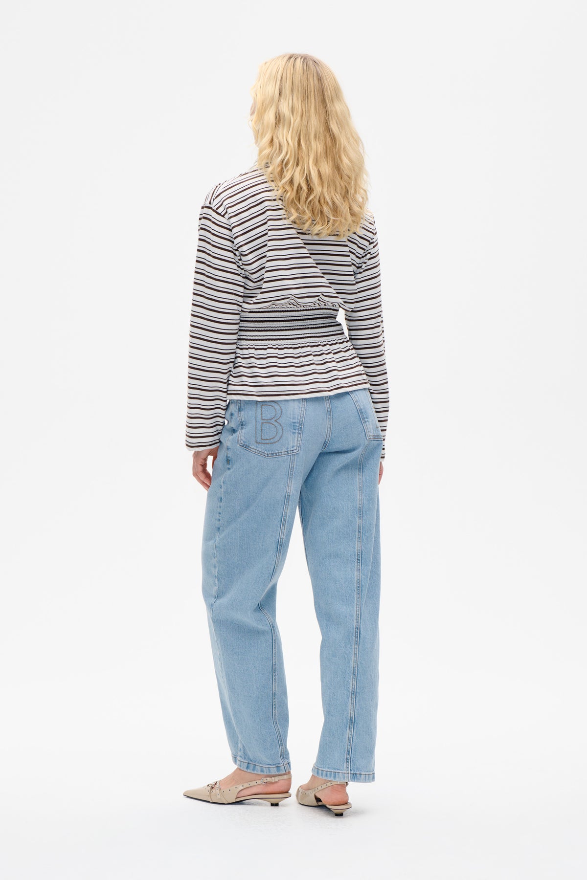 Back view of a model wearing a white long-sleeved top with narrow black stripes and a gathered waist, paired with light blue jeans.