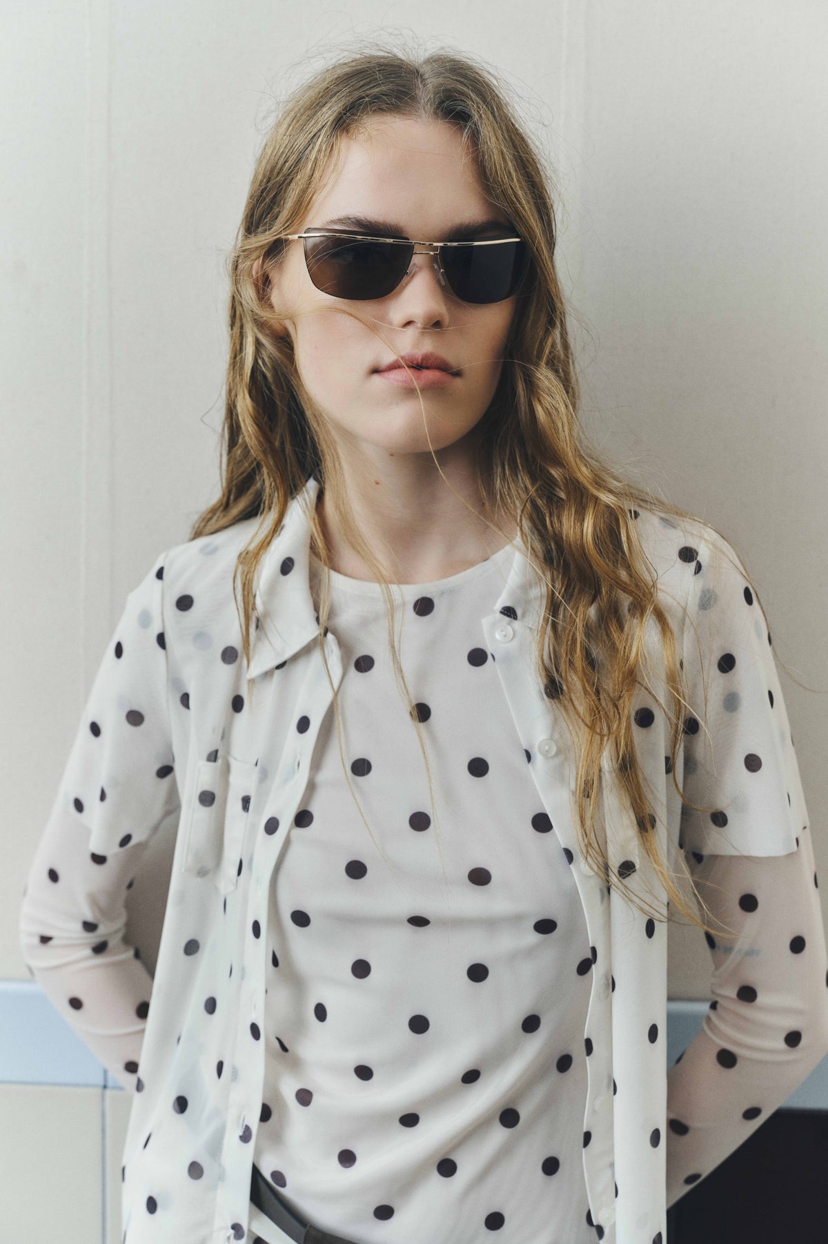 Close-up campaign portrait of a model wearing dark-lensed sunglasses and a white button-front shirt with black polka dots.