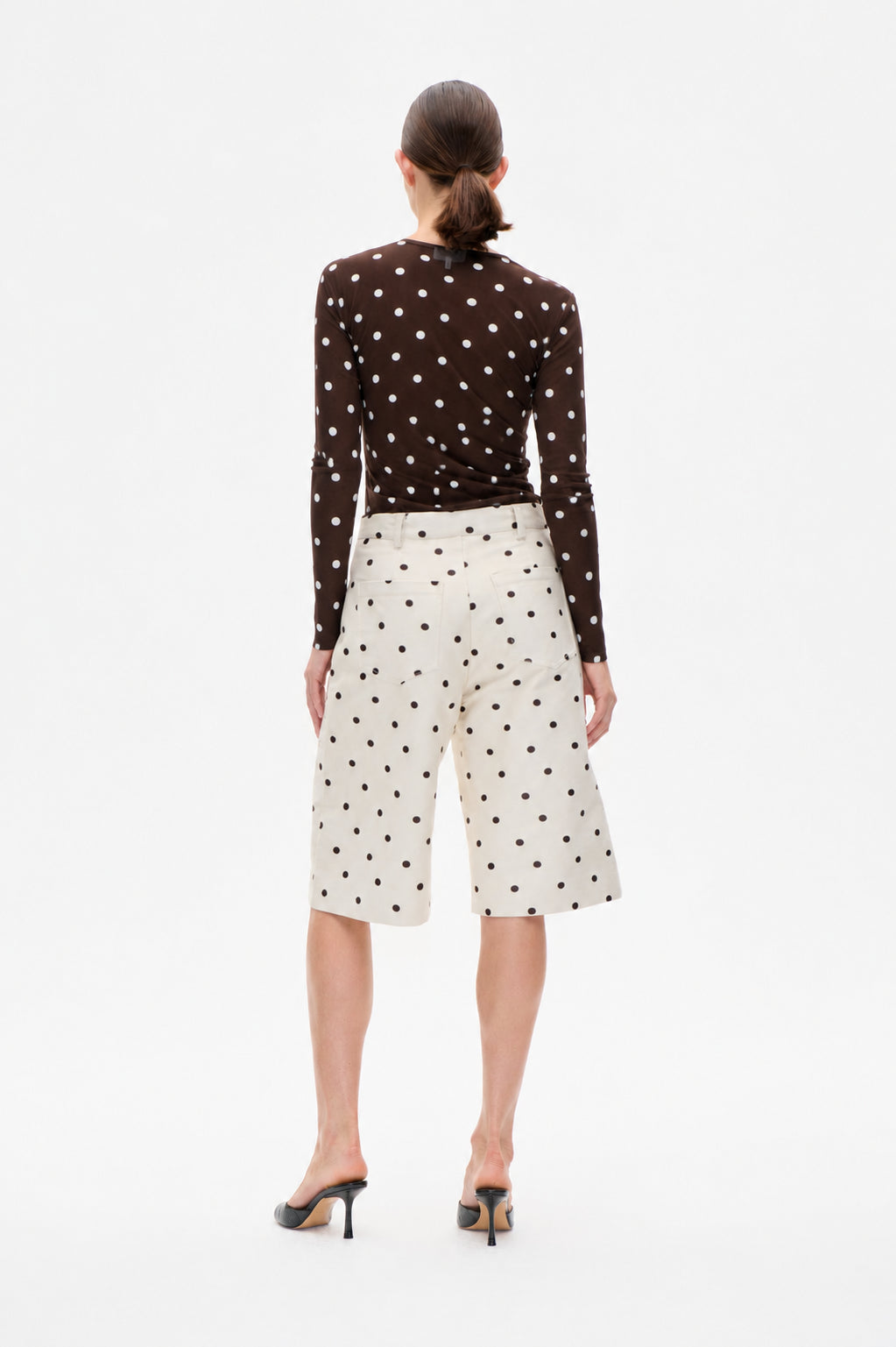 Back view of a model wearing a dark brown long-sleeve top with white polka dots and ivory knee-length shorts with dark polka dots.