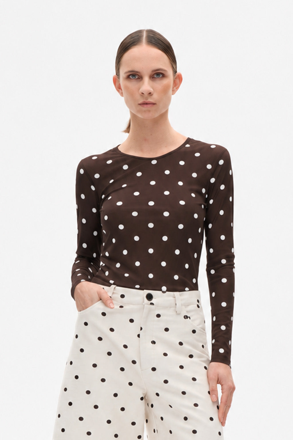 Model wearing a dark brown long-sleeve top with white polka dots and cream trousers with black polka dots.