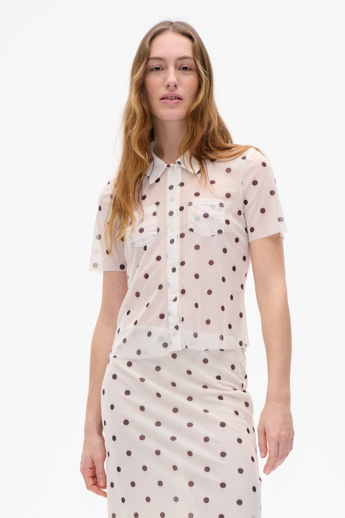 Cropped model wearing a cream short-sleeved button-front shirt with dark polka dots and chest pockets, paired with a matching skirt.