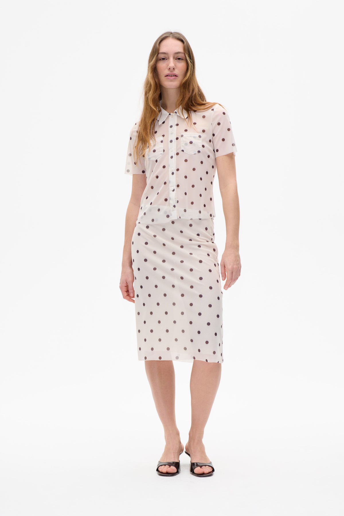 Model wearing a sheer pale pink short-sleeved button-front blouse and matching knee-length skirt with dark polka dots against a white backdrop.