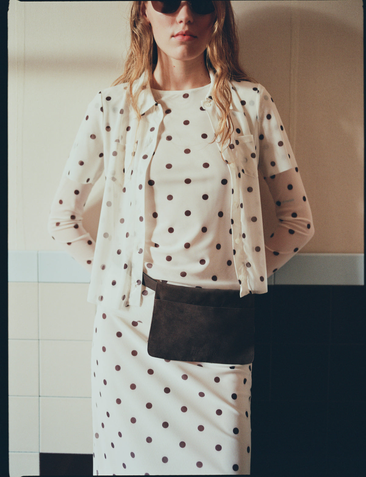 Cropped campaign portrait of a model in a cream outfit with dark polka dots, layering an open long-sleeved shirt over a top and skirt, with a dark shoulder bag.