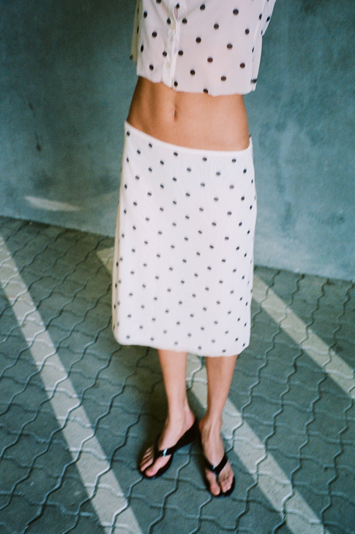 Cropped campaign view of a white black-polka-dot button-front shirt and matching knee-length skirt, with a bare midriff and black sandals.