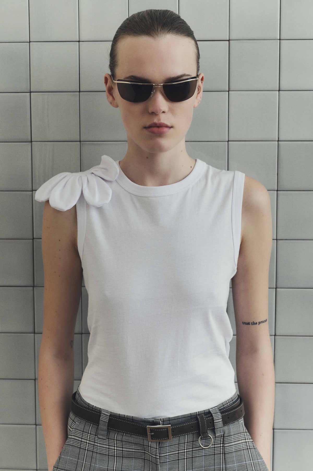 Front-facing campaign portrait in a white sleeveless top with a large shoulder bow, black-and-white plaid trousers, layered belts and dark sunglasses.