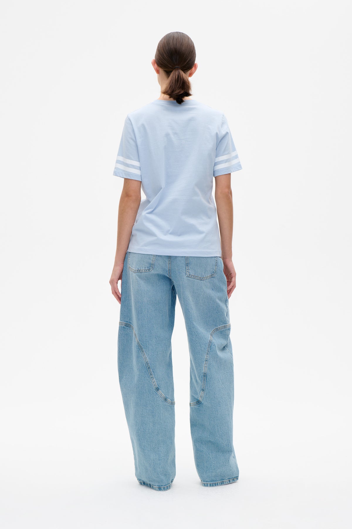Back view of a model wearing a pale blue short-sleeved T-shirt with two white stripes on each sleeve and wide-leg light-wash jeans.