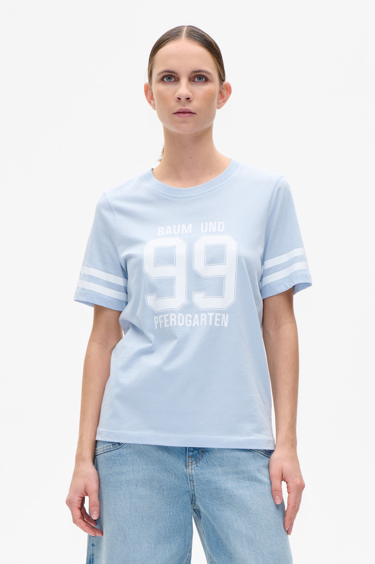 Front view of a pale blue short-sleeve T-shirt with white “BAUM UND PFERDGARTEN 99” lettering and striped sleeves, styled with light-wash jeans.