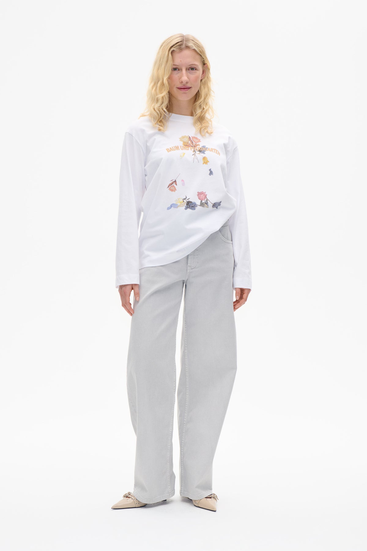 Model wearing a white long-sleeved T-shirt with a multicoloured floral front graphic and light grey wide-leg jeans.