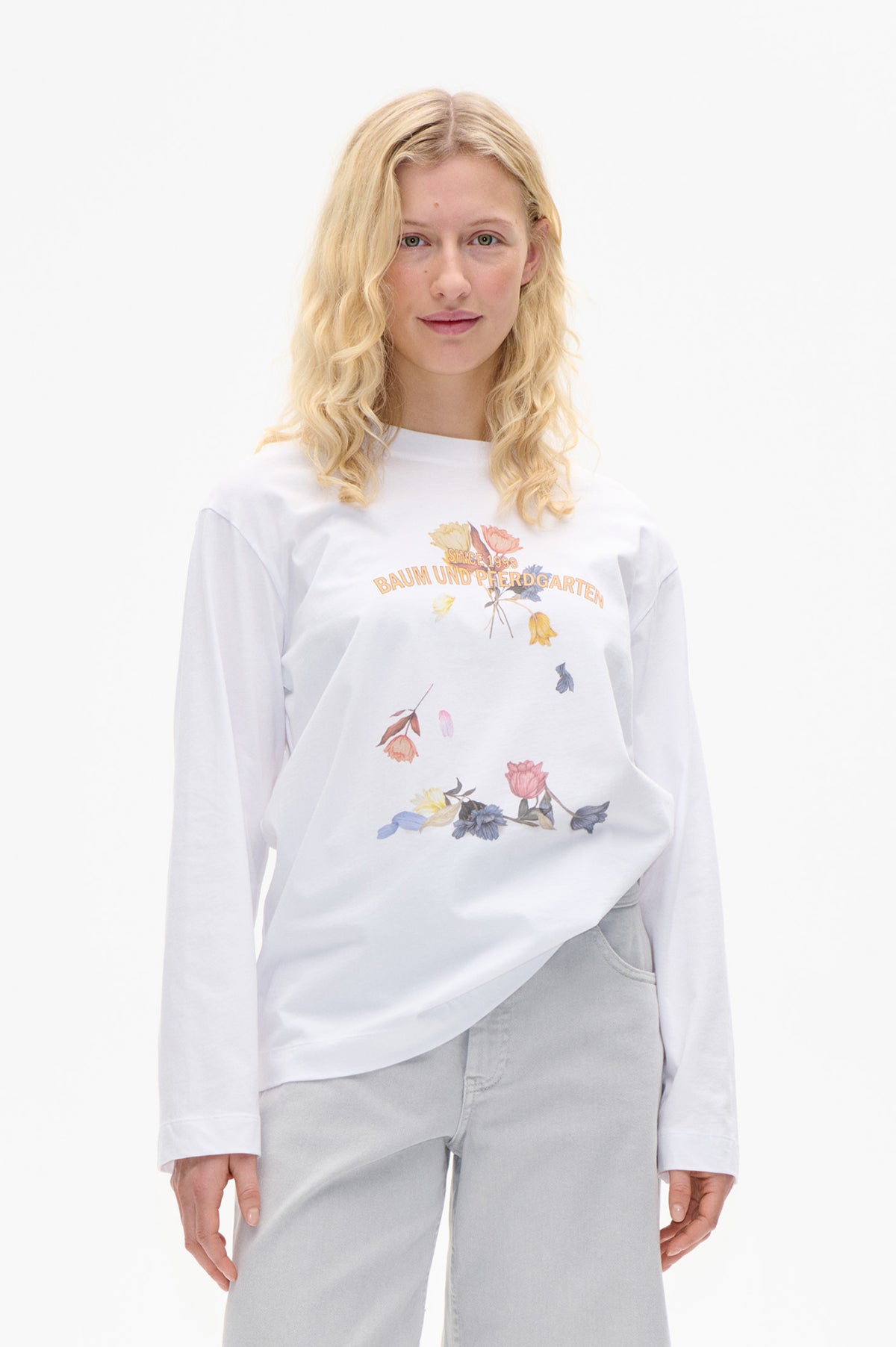 Model wearing a white long-sleeved T-shirt with colorful floral graphics and “Since 1999 Baum und Pferdgarten” lettering, shown from the front.