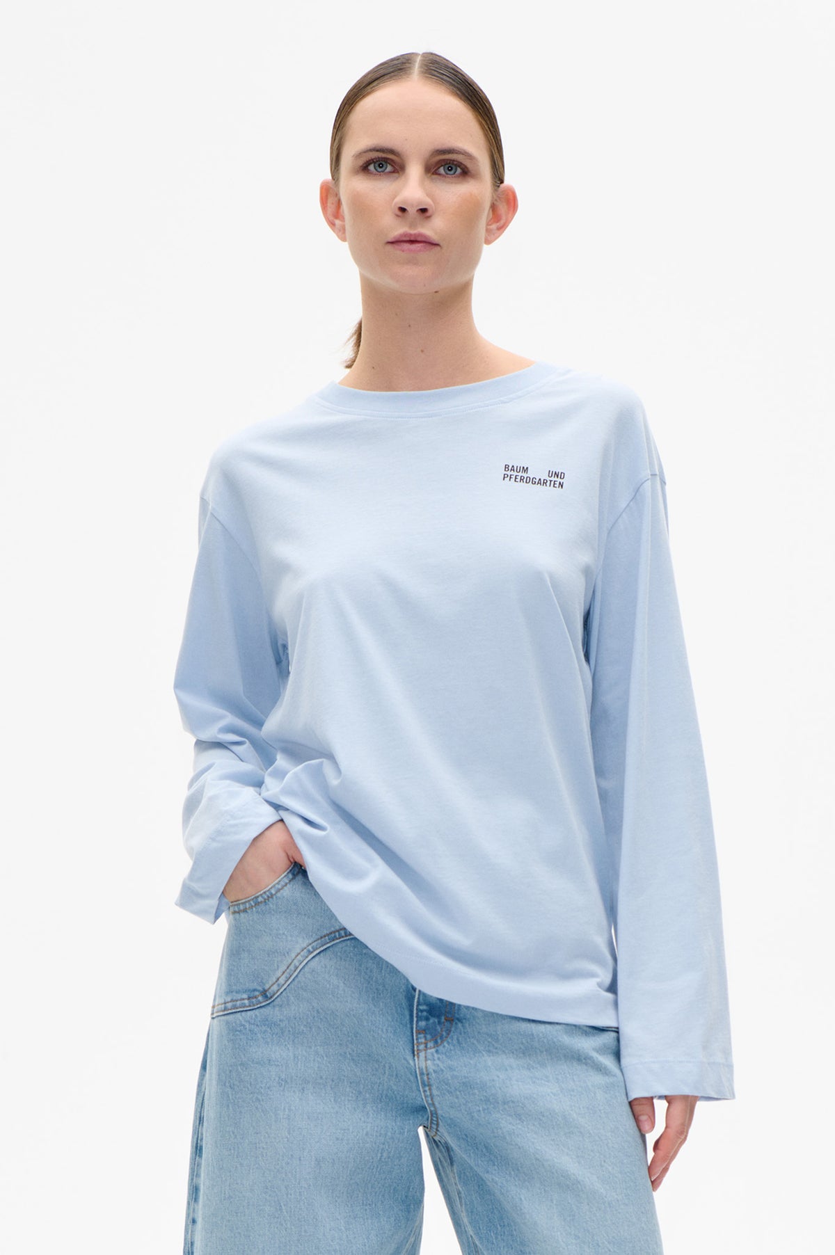 Model wearing a pale blue oversized long-sleeve sweatshirt with small Baum und Pferdgarten chest lettering and light-wash jeans, front view.