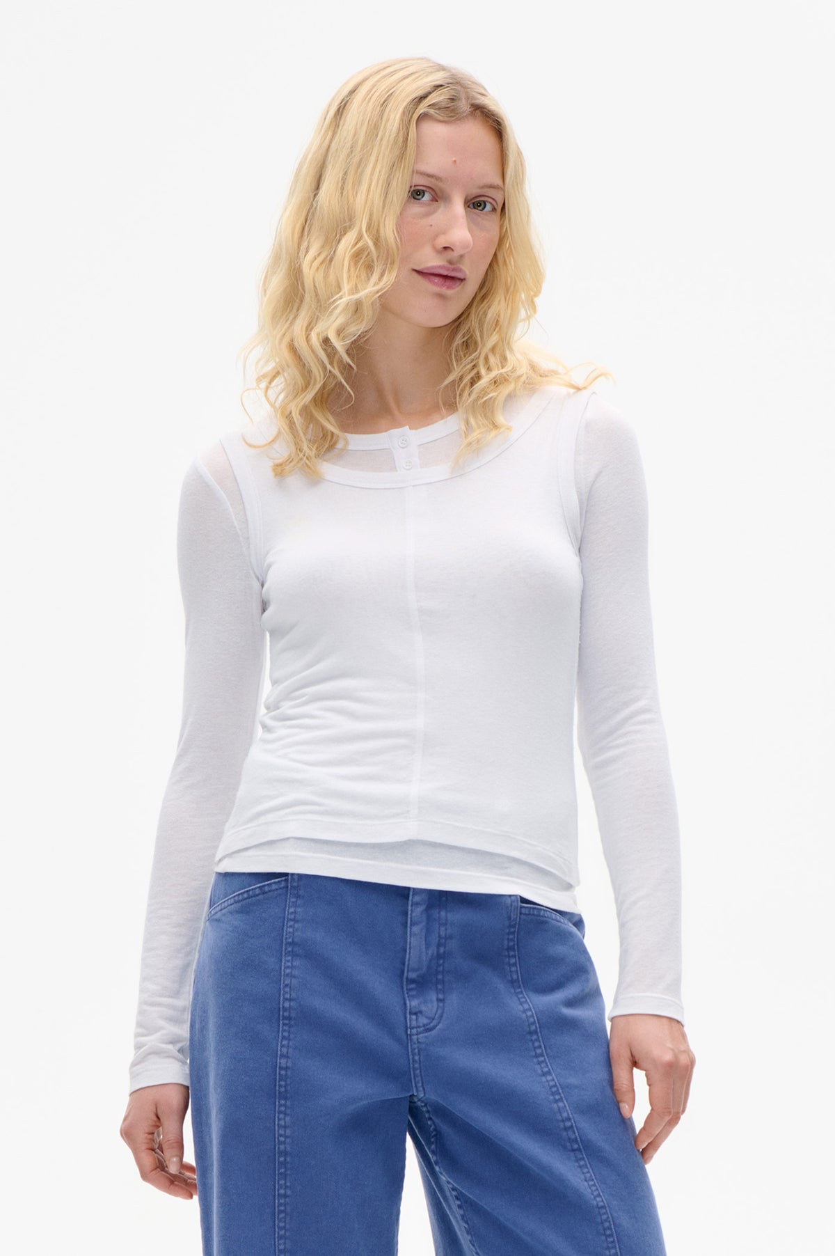 Vorderansicht eines Models, das ein weißes Langarmtop mit geschwungenem Lagen-Ausschnitt und kleinen Knöpfen trägt, kombiniert mit blauen Jeans.