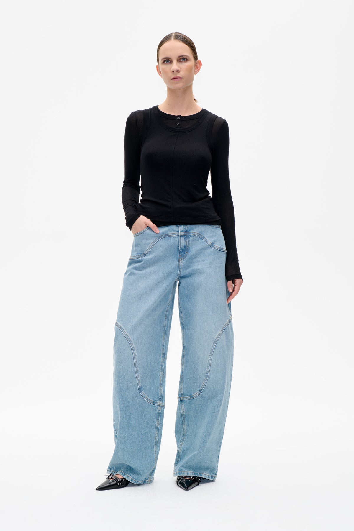 Vorderansicht eines Models in schwarzem, mehrlagigem Top mit transparenten Ärmeln und knöpfbarem Ausschnitt, kombiniert mit hellblauen, weit geschnittenen Jeans.