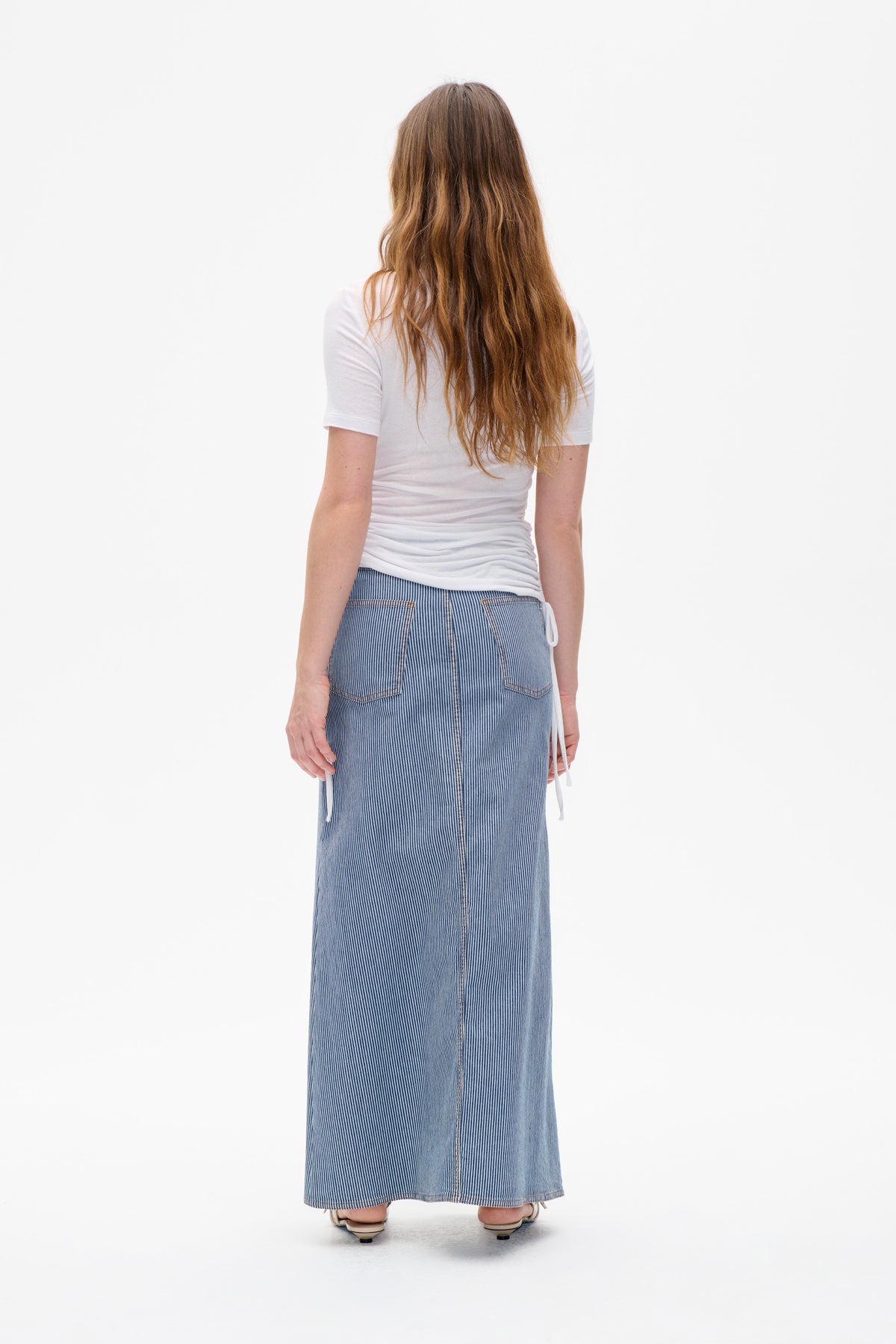 Back view of a model wearing a plain white short-sleeved T-shirt and a long blue-and-white pinstriped skirt with patch pockets and side ties.
