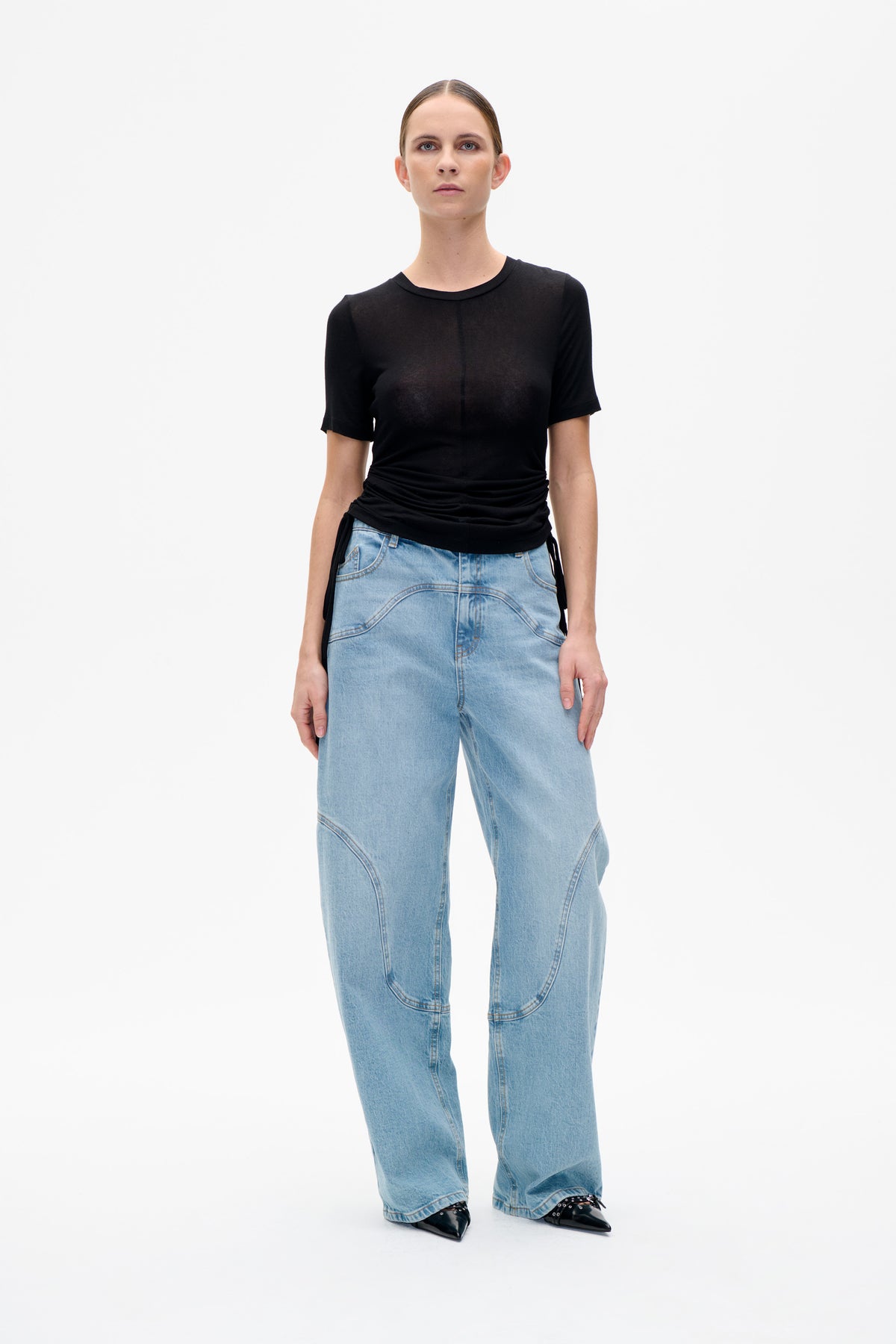 Frontansicht eines Models in einem schwarzen, kurzärmeligen Rundhals-Top mit gerafftem Saum und Bindebanddetail sowie einer hellgewaschenen, weit geschnittenen Jeans.