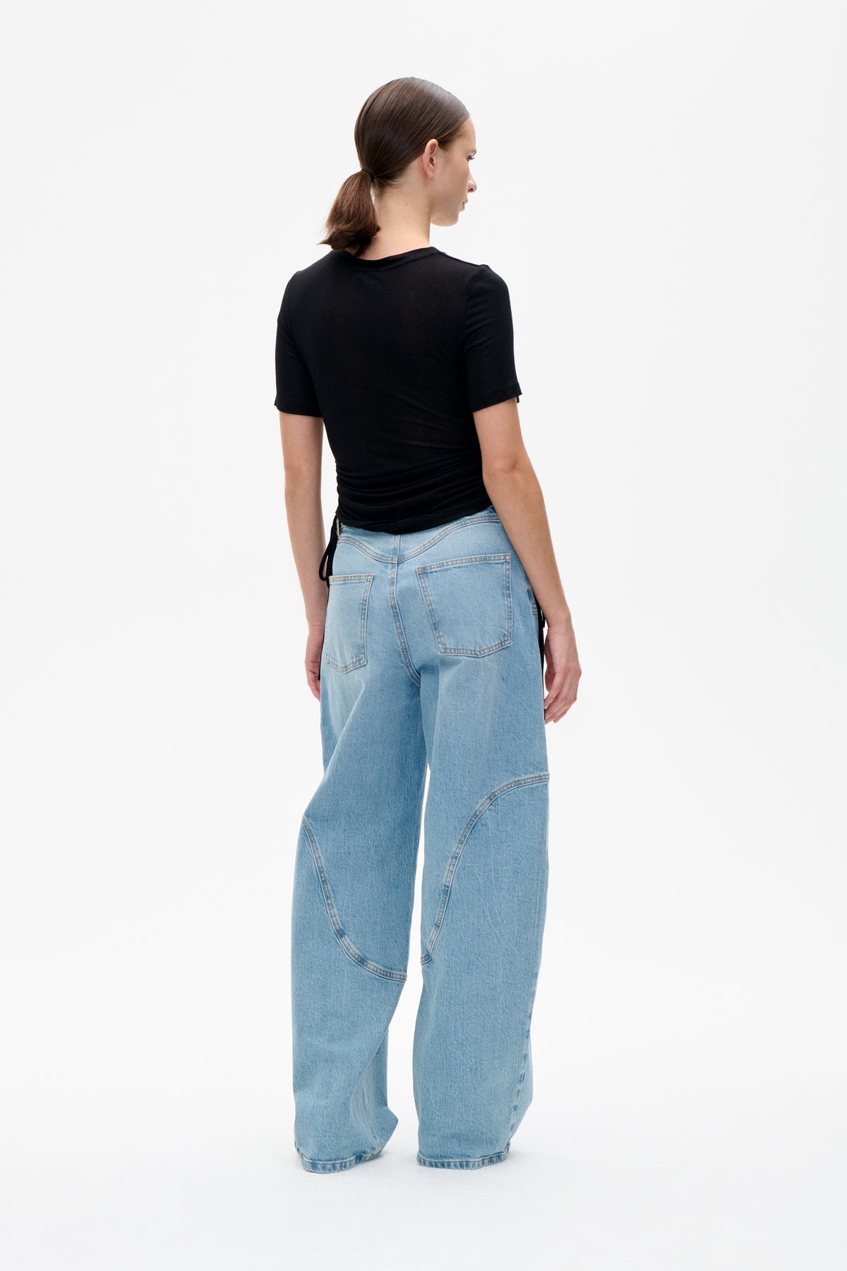 Rückansicht eines Models in einem figurbetonten schwarzen Kurzarmtop mit seitlichen Bindebändern und hellblau gewaschenen, weit geschnittenen Jeans.