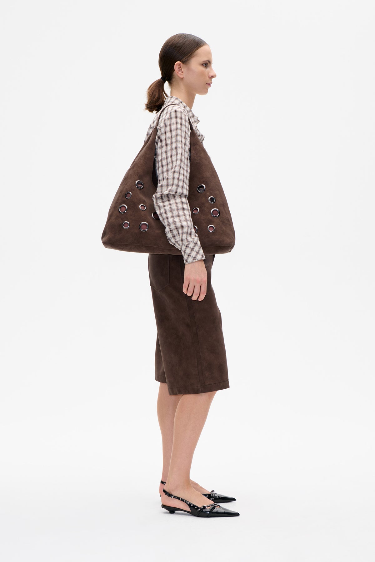 Side-profile studio styling of a large brown tote with metal eyelets, a cream plaid ruffle-neck blouse, brown knee-length bottoms and black flats.