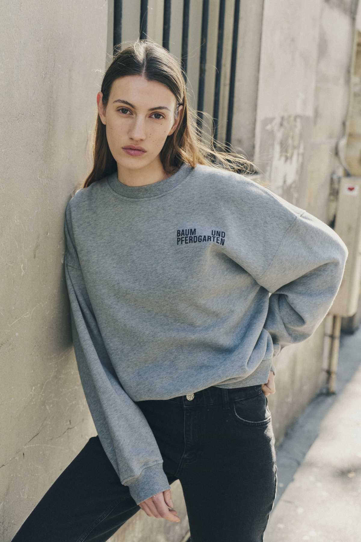 Model wearing a light grey oversized sweatshirt with a dark Baum und Pferdgarten chest logo and black jeans, leaning against a wall.