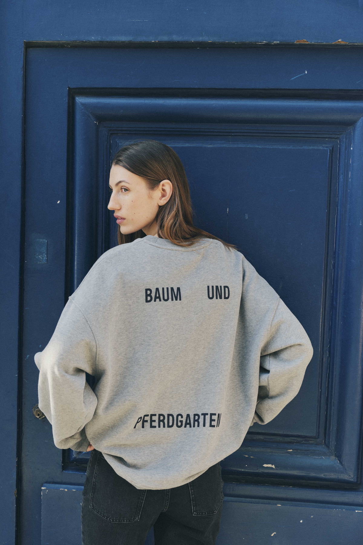 Model wearing a loose grey crewneck sweatshirt with black Baum und Pferdgarten lettering across the back, against a deep blue door.
