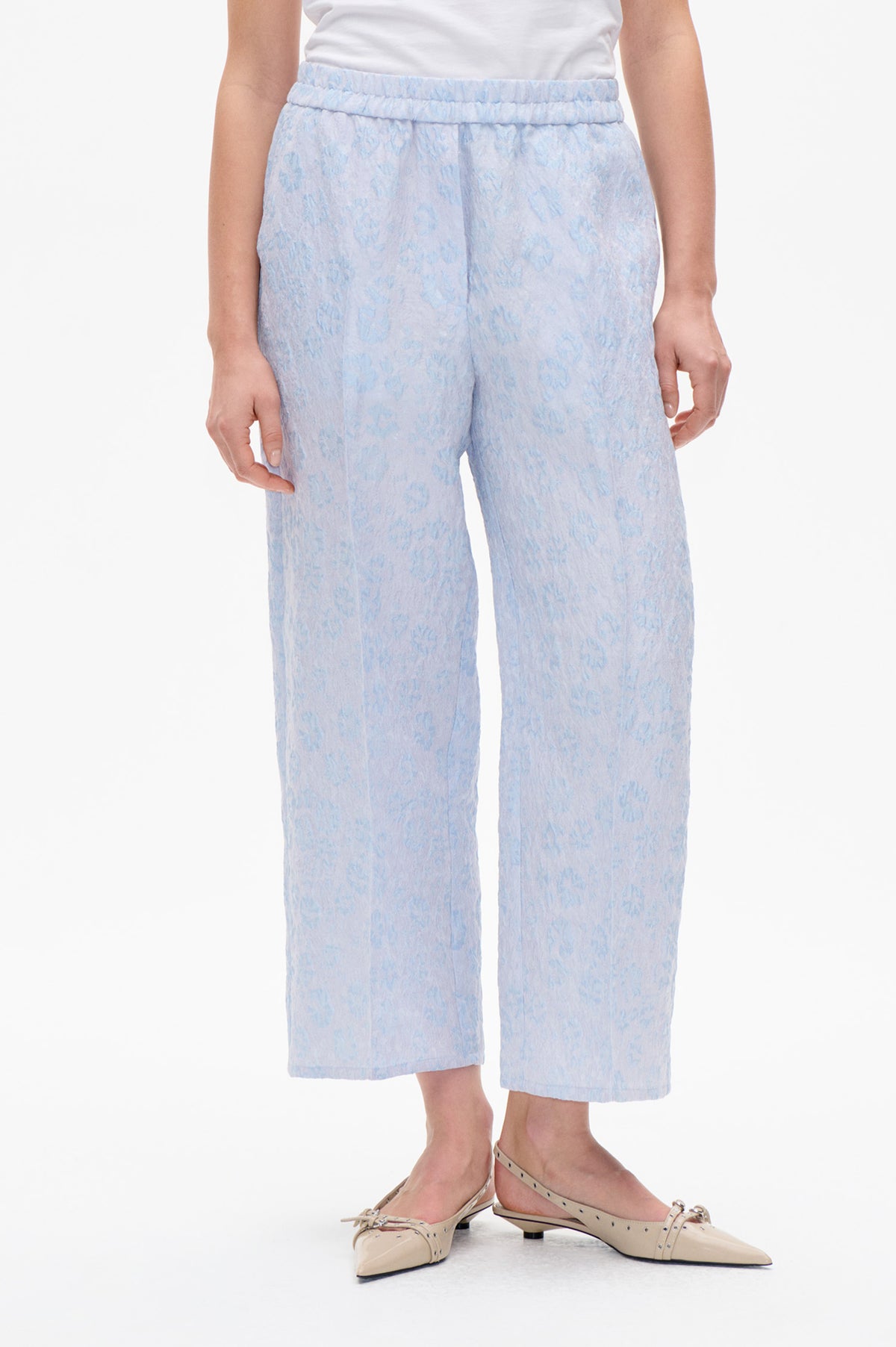 Front detail of light blue cropped trousers with a gathered elastic waistband and subtle tonal floral pattern, styled with a white top.