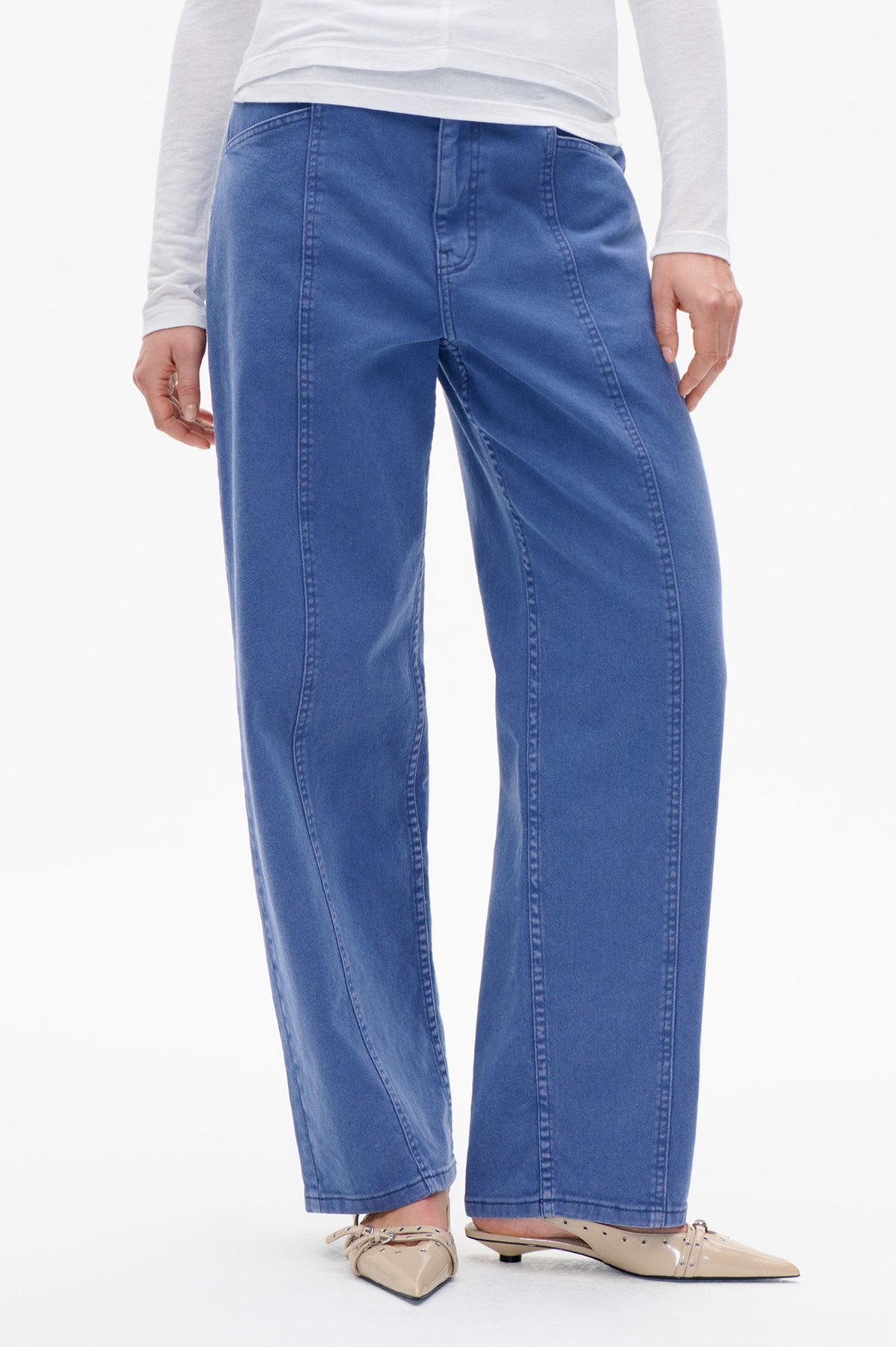 Close front view of blue wide-leg jeans with curved front pockets and prominent vertical panel seams, styled with a white top.