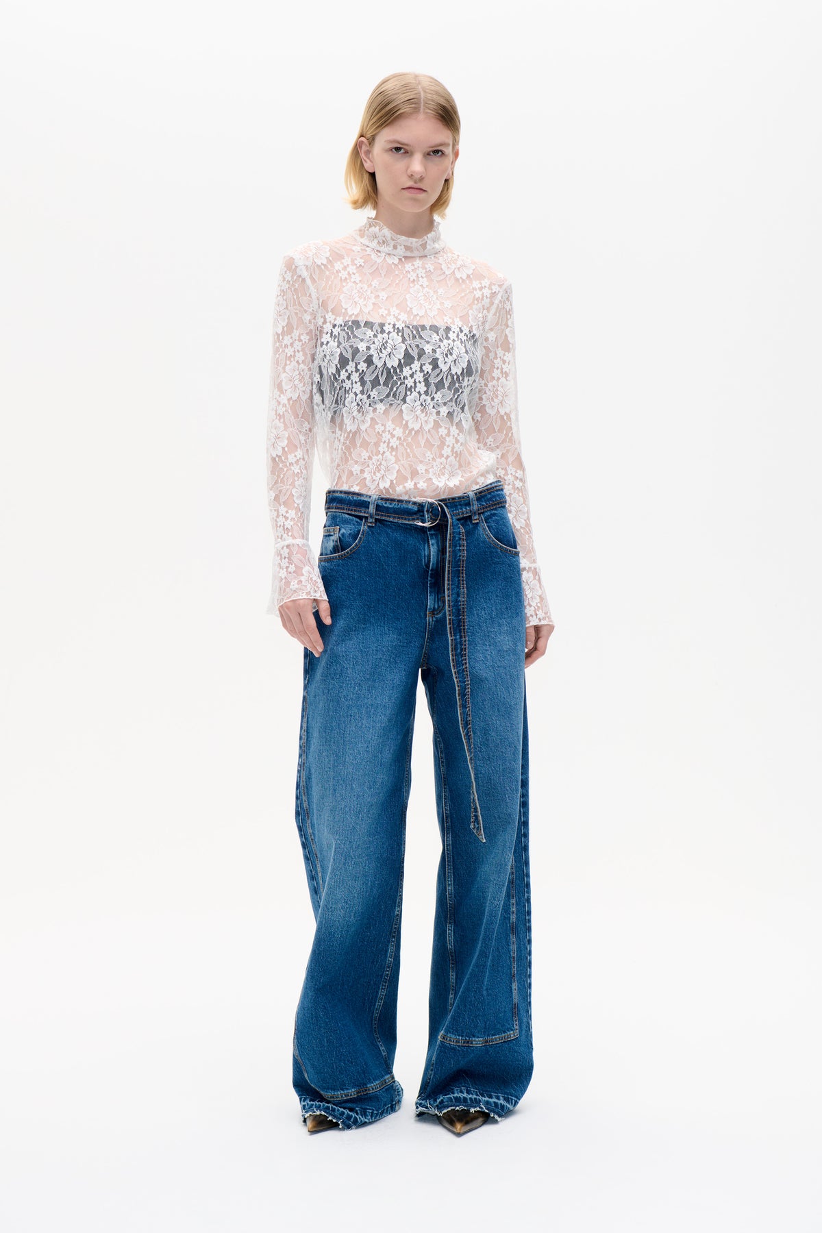 Front view of a model wearing a sheer white floral lace long-sleeved top with a high neck and wide-leg blue jeans.