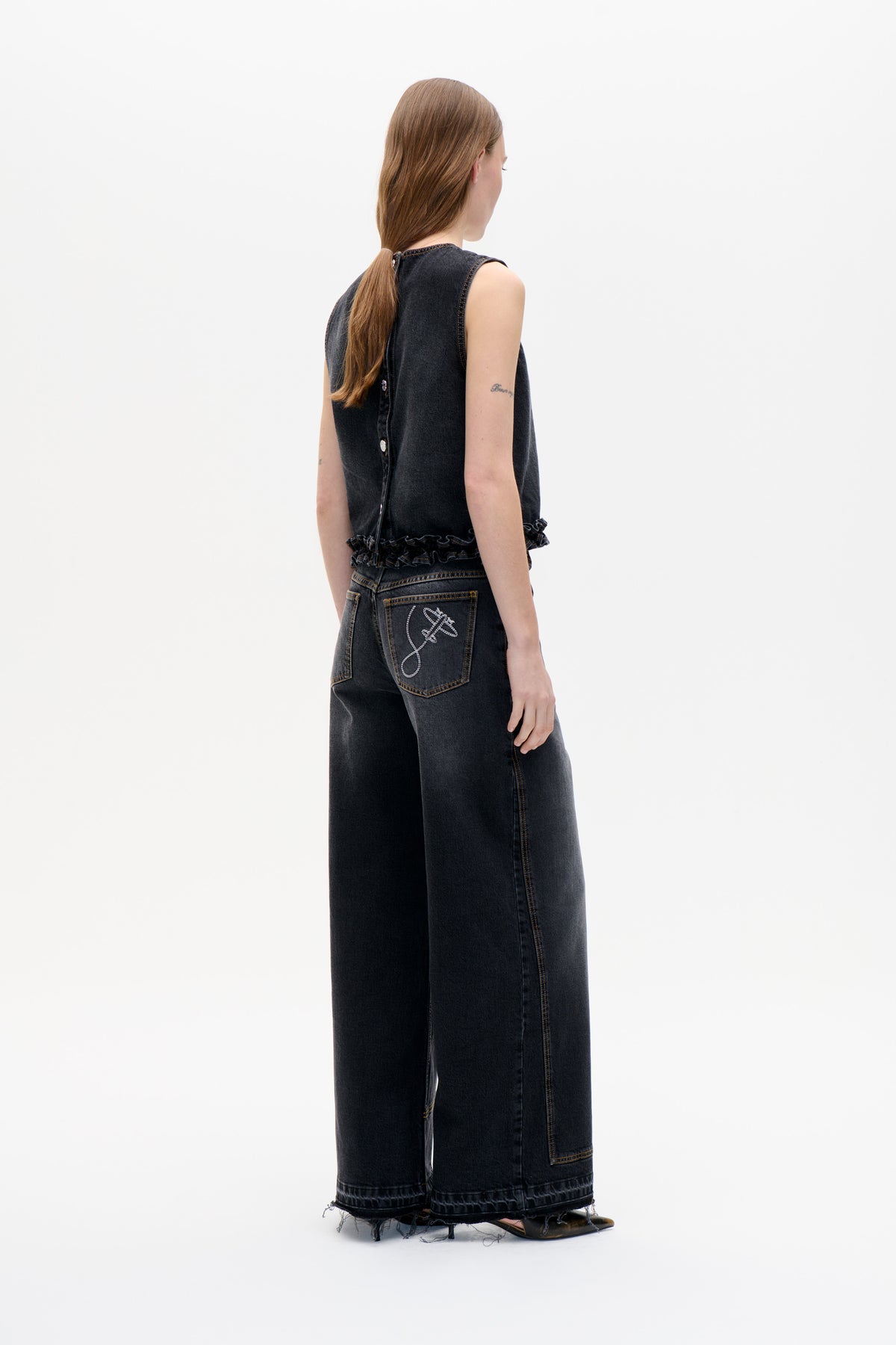 Model shown from behind in a charcoal sleeveless denim top with a ruffled hem and wide-leg jeans with embroidered back pocket and frayed hems.