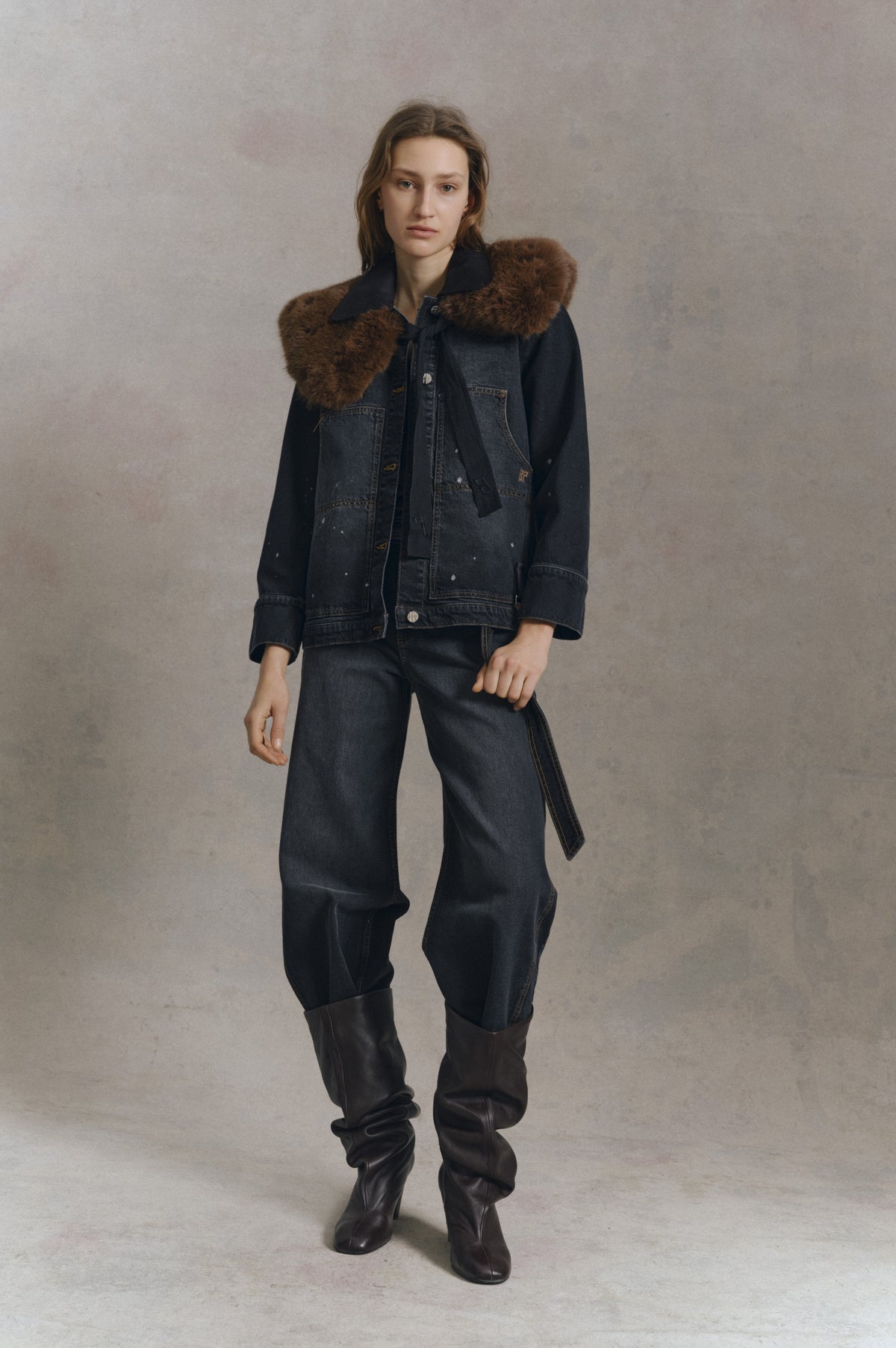 Model wearing a dark denim jacket with brown fur trim and matching wide-leg jeans, styled with tall dark boots against a neutral backdrop.