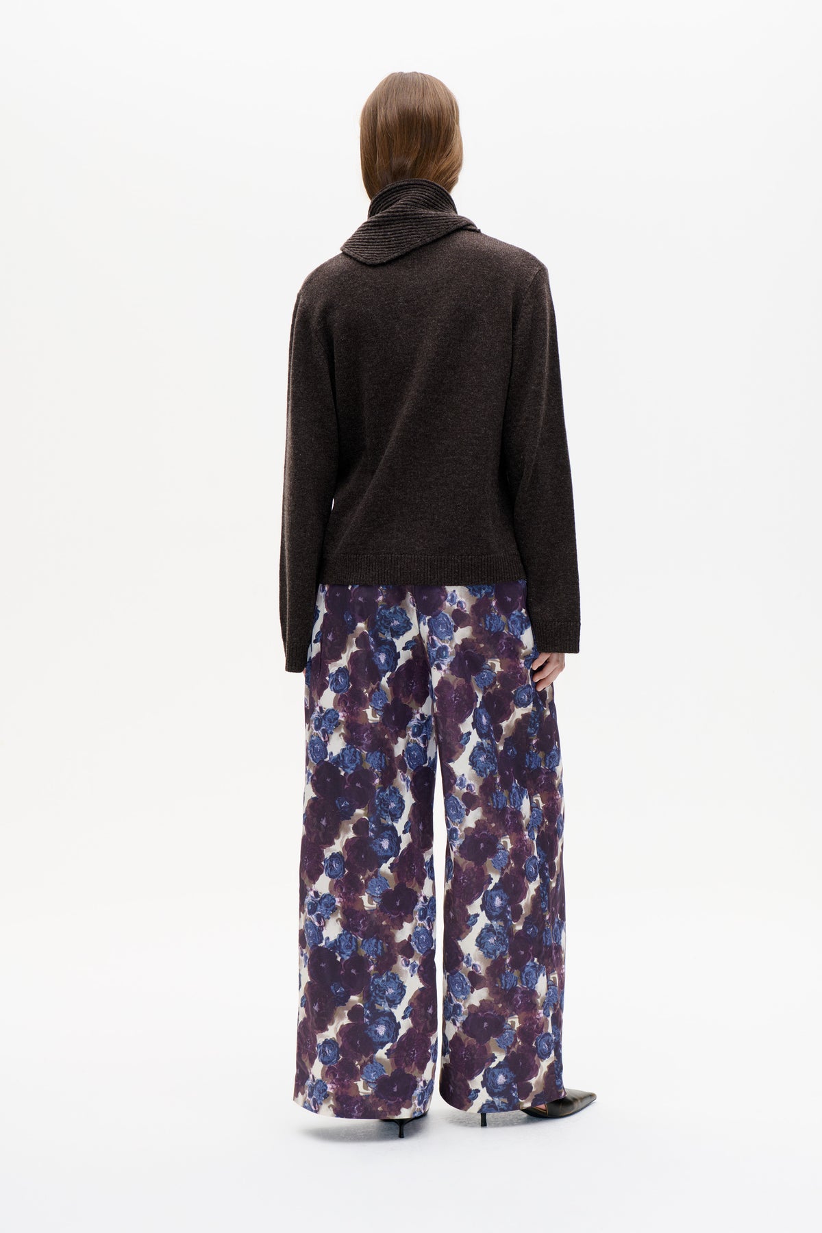 Back view of a model in a dark charcoal-brown sweater with a ribbed collar and wide-leg cream trousers printed with blue and purple flowers.