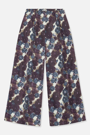 Vooraanzicht van een broek met wijde pijpen, knoopsluiting in de taille, plooien en grote bloemen in pruim en blauw op een crèmekleurige achtergrond