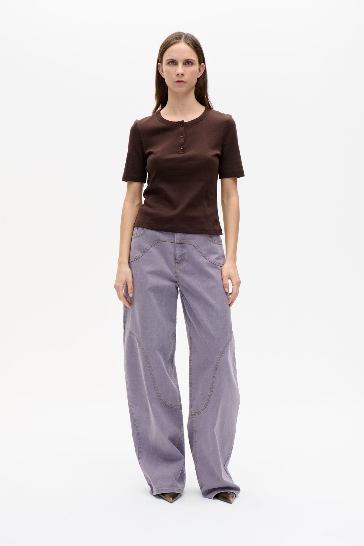 Front view of a model wearing a dark brown short-sleeved henley top with three buttons and lilac wide-leg jeans.