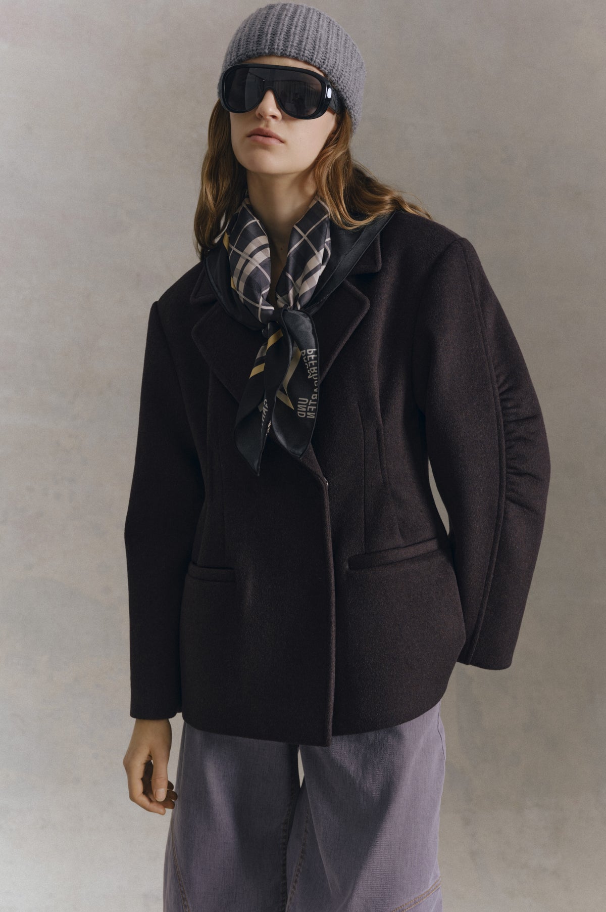 Model wearing a dark plum coat, grey ribbed beanie, oversized black sunglasses and a grey checked scarf, with lilac trousers.