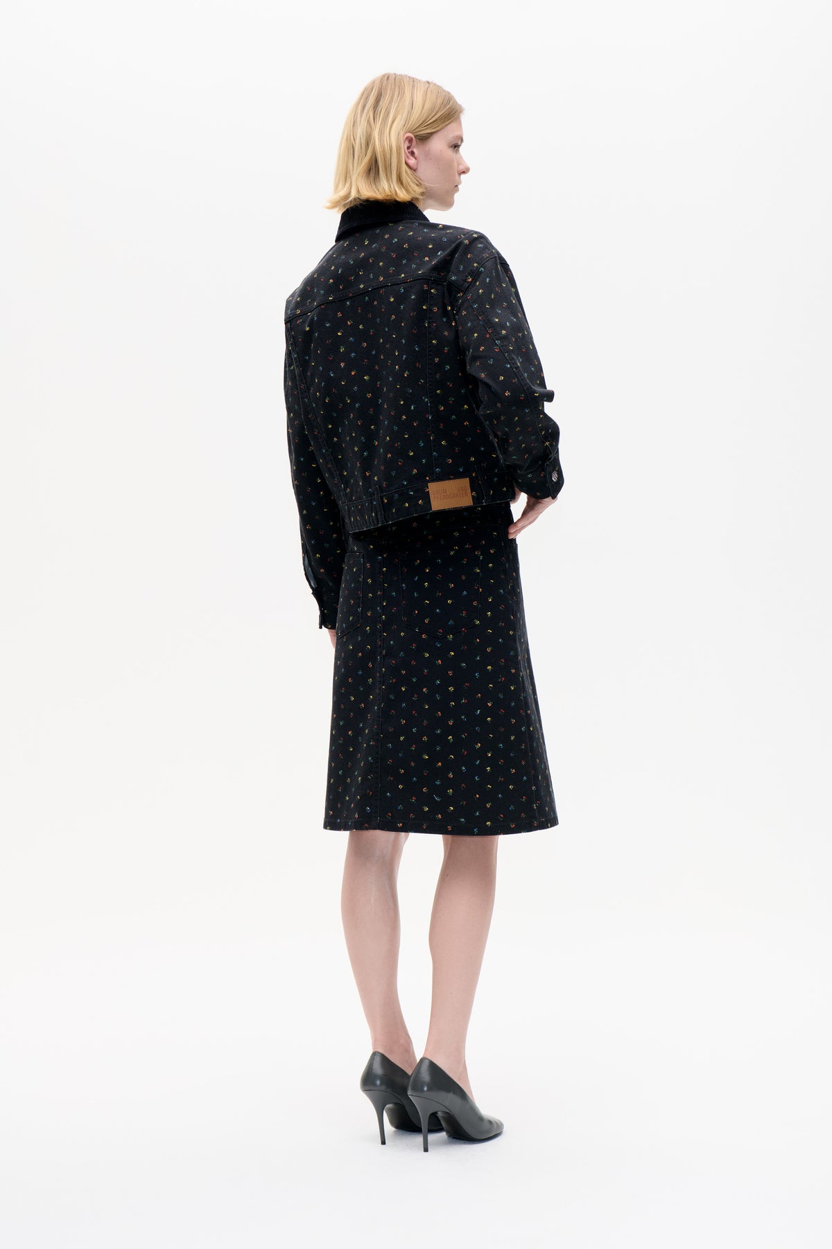 Model viewed from behind wearing a black jacket and matching knee-length skirt with scattered multicoloured floral specks.
