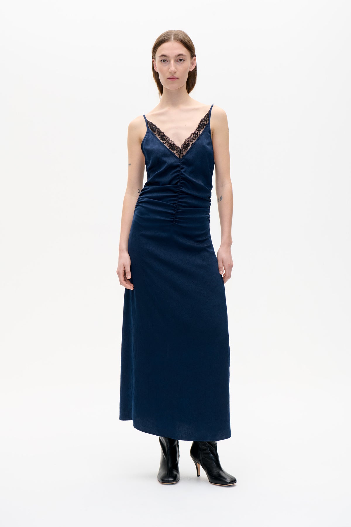 Front view of a model wearing a dark navy sleeveless dress with a V-neck trimmed in black floral lace and a gathered front seam.