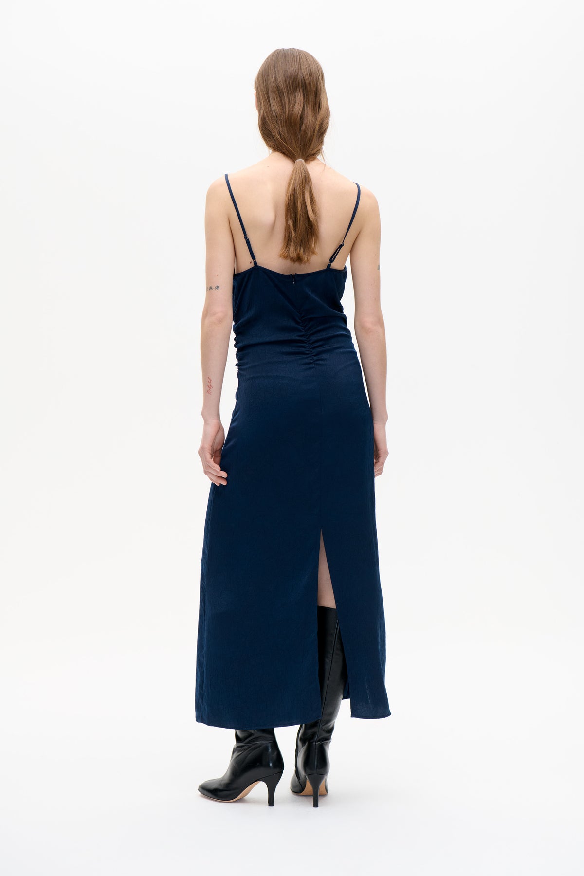 Back view of a model wearing a long navy spaghetti-strap dress with adjustable straps, ruched sides and a back slit.