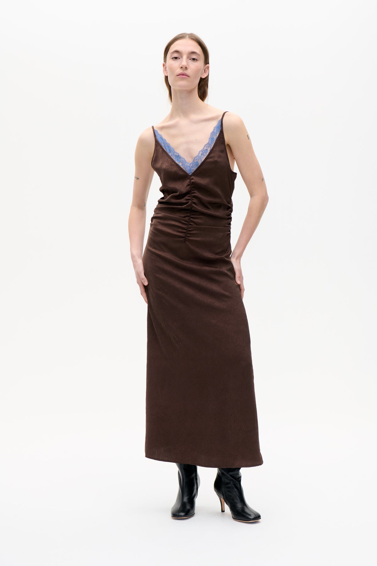 Front view of a model wearing a dark brown sleeveless midi dress with a ruched center seam and blue lace trim at the V-neck.