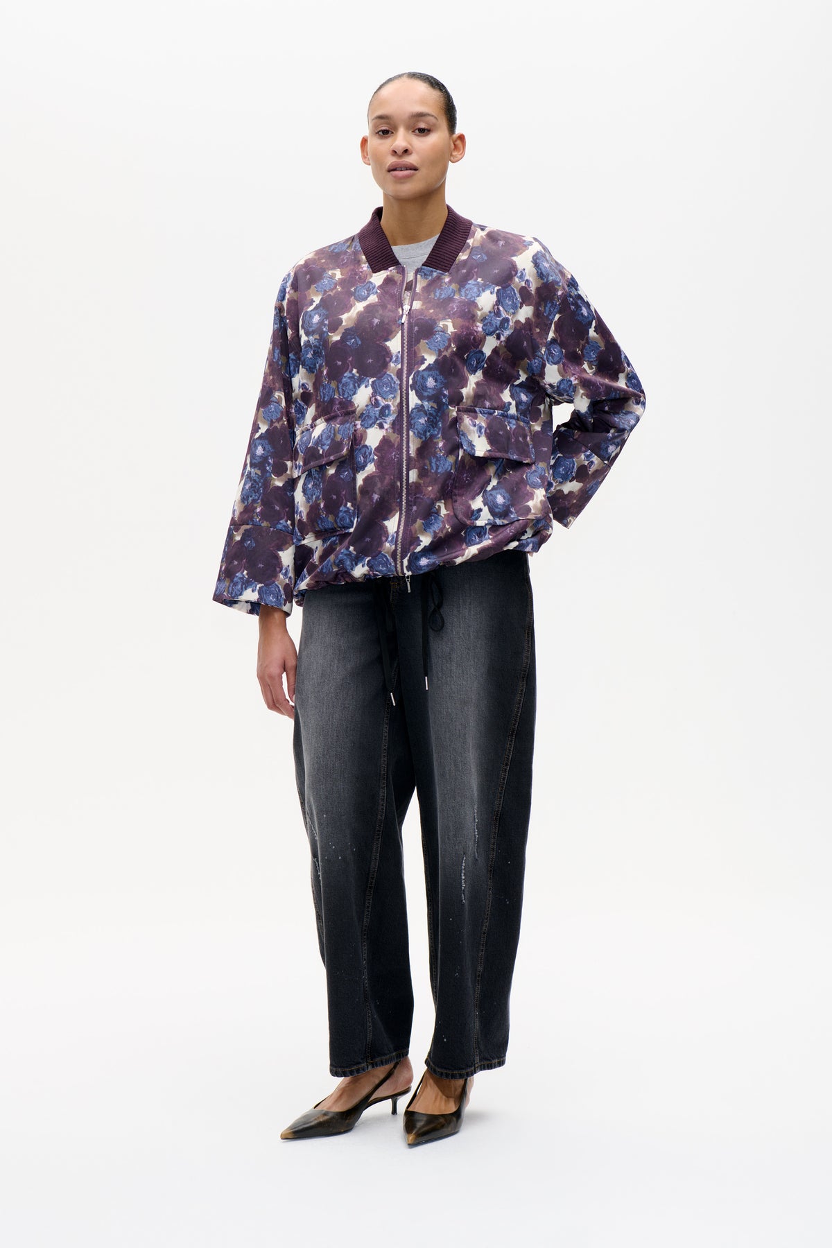 Front view of a model wearing a zip-front bomber jacket with a plum and blue floral print, ribbed collar and flap pockets.